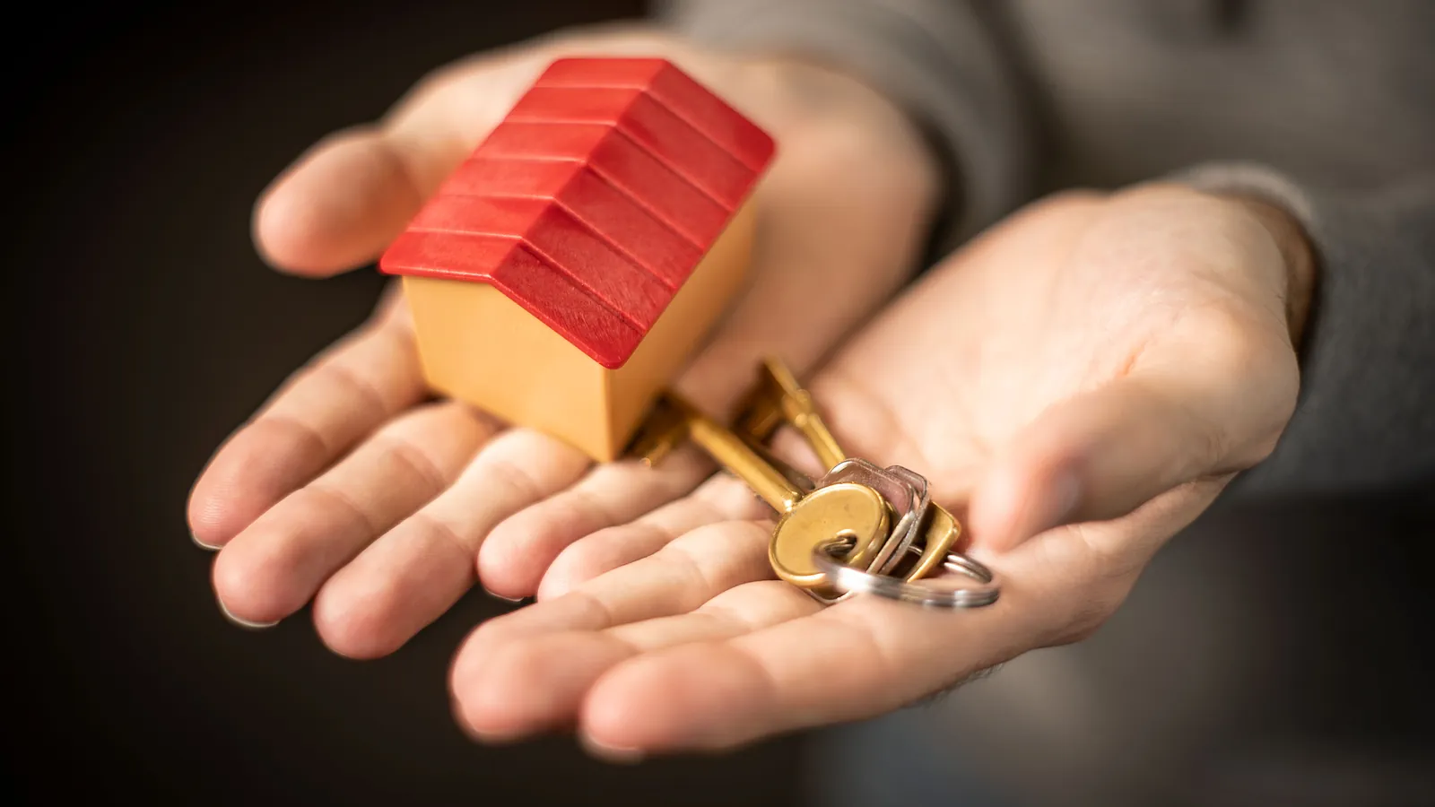 A person holding out a small model house in one hand and a set of house keys in the other, ready to hand a rented home to its tenant