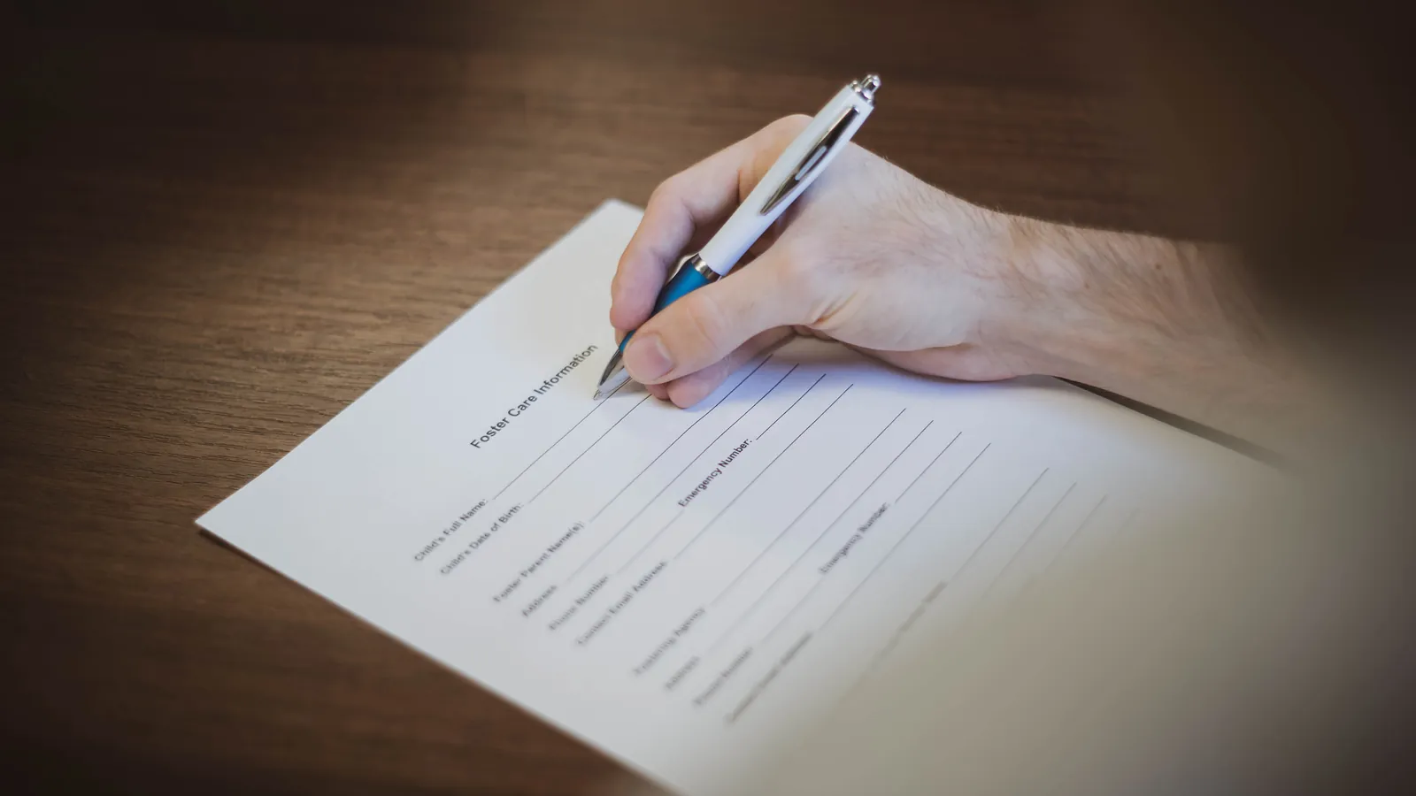 A hand filling in a paper form with a pen on a wooden desk